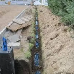 Długi wykop w ziemi z widocznymi kablami w niebieskich rurach ochronnych, ułożonymi wzdłuż wykopu. W tle widoczny betonowy fundament i fragment zieleni.