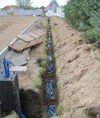 Długi wykop w ziemi z widocznymi kablami w niebieskich rurach ochronnych, ułożonymi wzdłuż wykopu. W tle widoczny betonowy fundament i fragment zieleni.