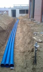 Rów z trzema niebieskimi rurami ułożonymi wzdłuż wykopu, widoczny fragment budynku w tle, ziemia piaszczysta.