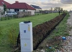 Biała skrzynka elektryczna na tle wykopu w ziemi, widoczny trawnik i domy w oddali, pochmurne niebo.