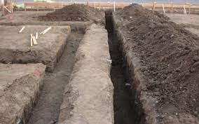 Wykopane wąskie rowy pod fundamenty, widoczne hałdy ziemi po bokach, kilka desek na ziemi, teren budowy w trakcie przygotowań.