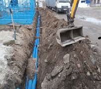 Wykopany wąski rów w ziemi z ułożonymi niebieskimi rurami, widoczna łyżka koparki i fragment ciężarówki w tle.