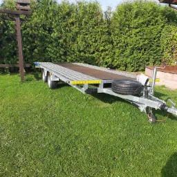 Płaska przyczepa samochodowa z zapasowym kołem, stojąca na trawniku przed żywopłotem z tui.