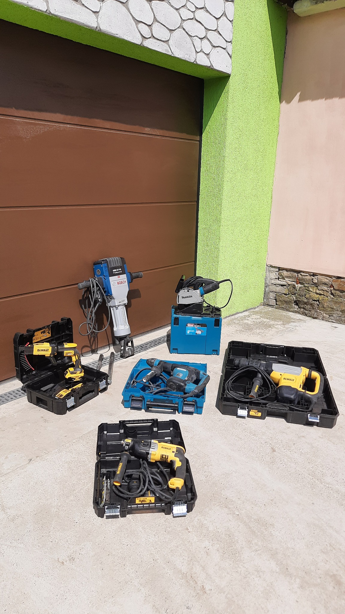 Zestaw sprzętu budowlanego różnych marek (Bosch, Makita, DeWalt) w otwartych skrzynkach na tle brązowej bramy garażowej i zielonej ściany.