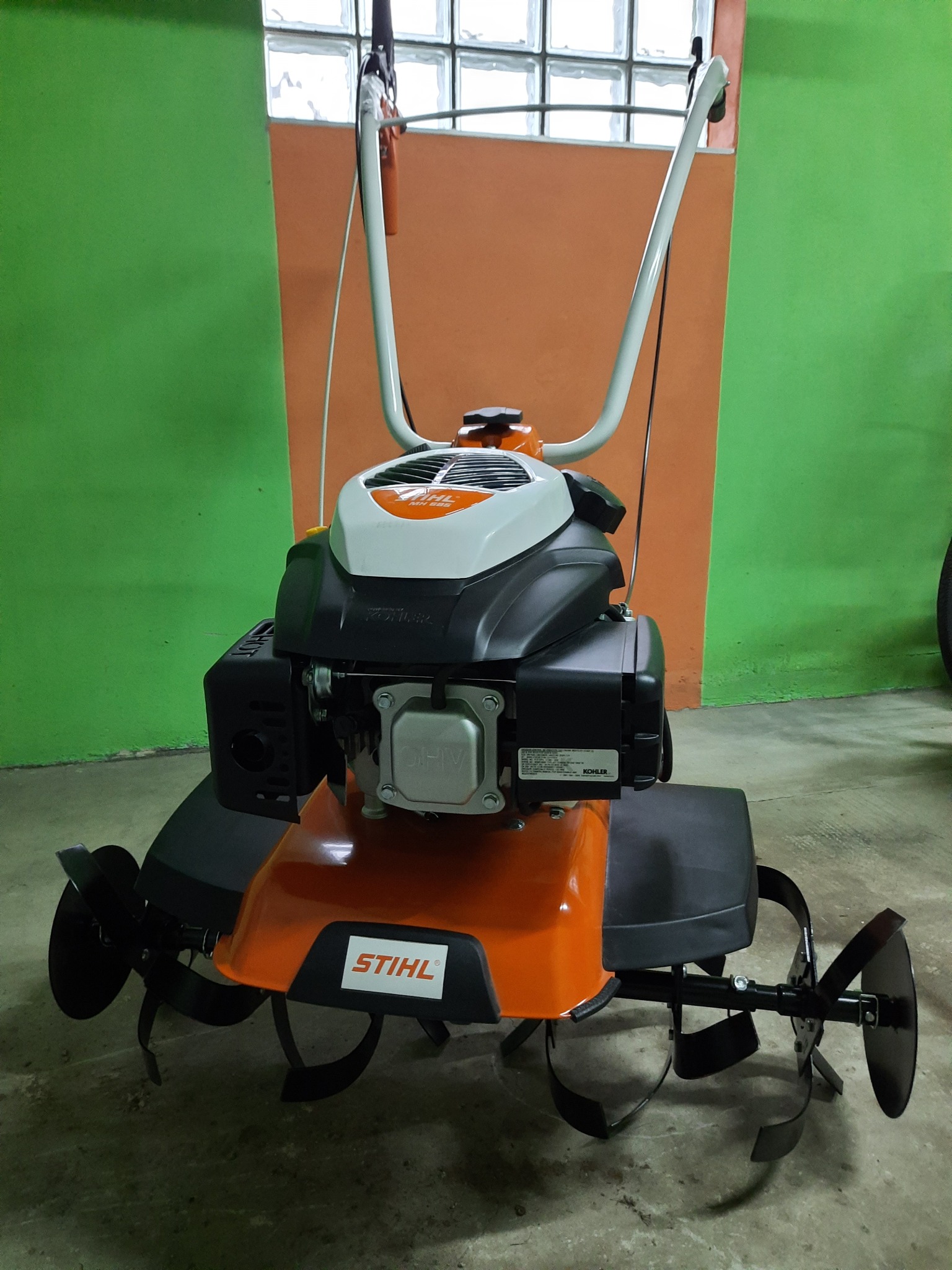 Stojący kultywator spalinowy Stihl MH 585 z widocznym silnikiem Kohler, na betonowej posadzce przed zielono-pomarańczową ścianą z luksferami.