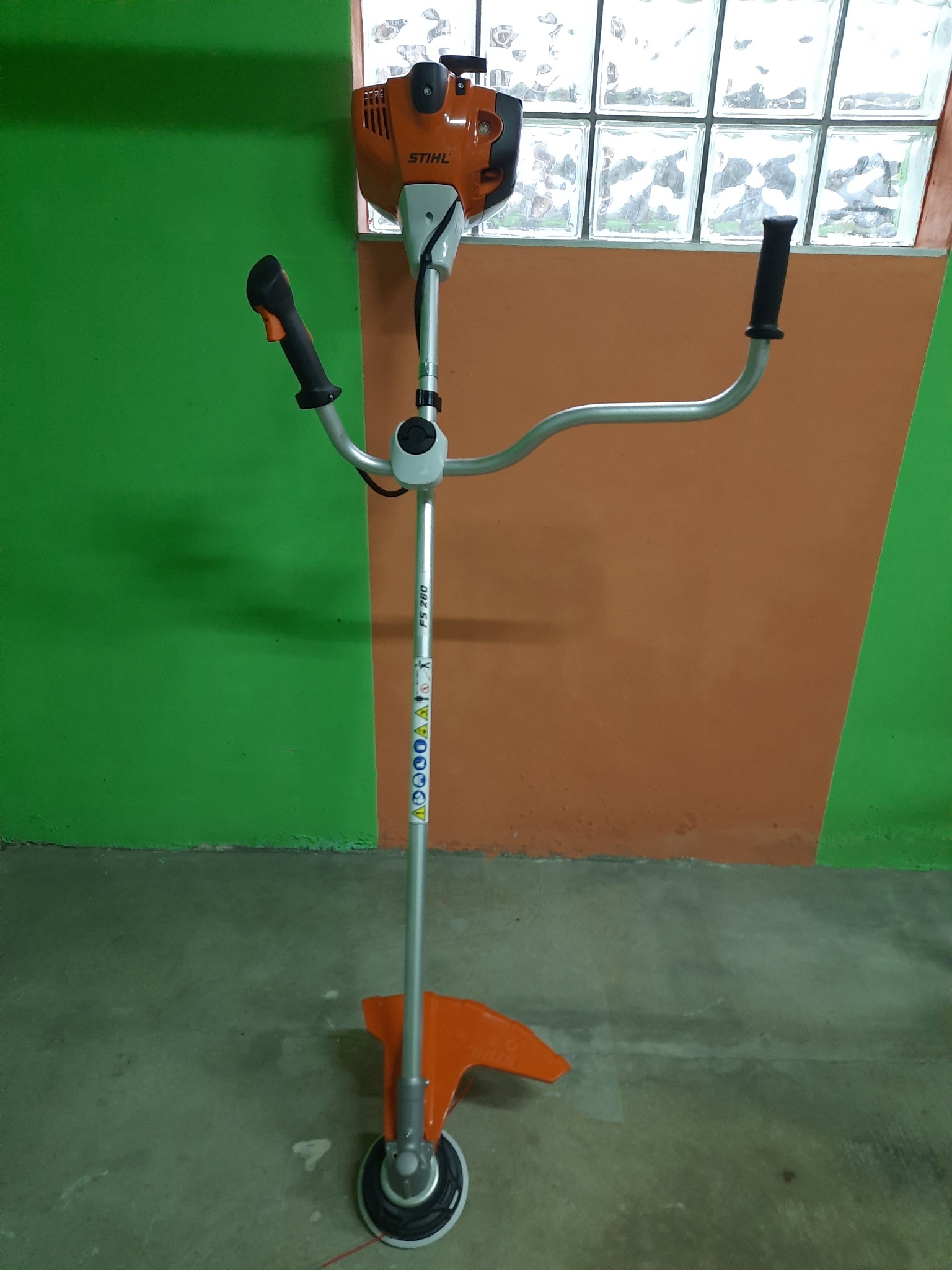 Pomarańczowo-srebrna kosa spalinowa Stihl FS 260, stojąca na szarym betonie, z zielono-pomarańczowym tłem i kratką okienną z luksferów.