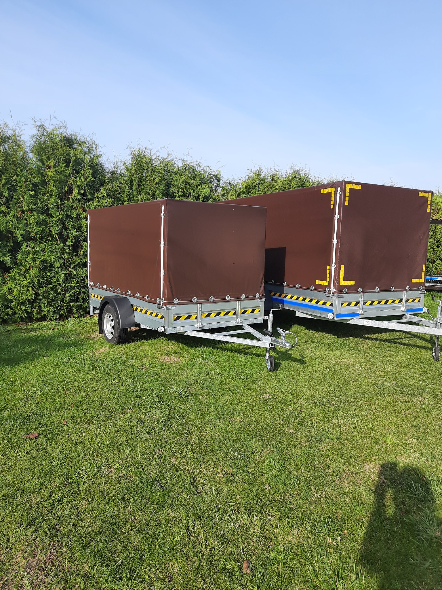 Dwie przyczepy budowlane z brązowymi plandekami, stojące na trawie w Mirzec, z widocznymi elementami odblaskowymi i zielonymi krzewami w tle.