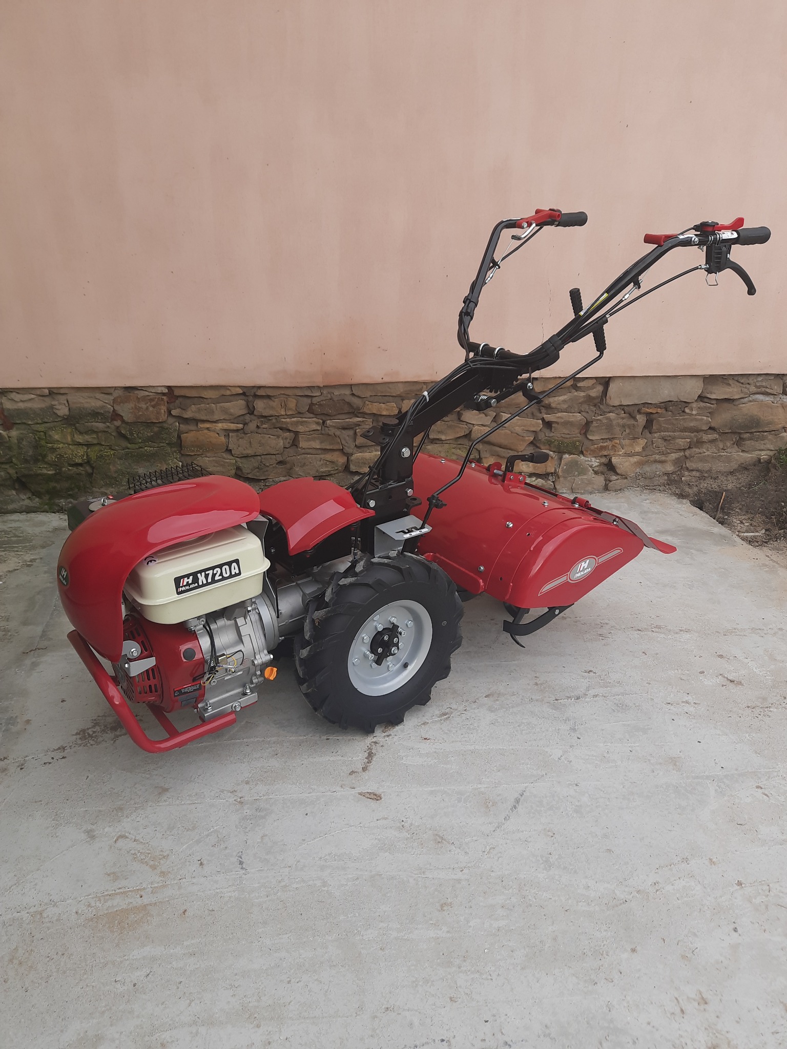 Czerwony glebogryzarek marki IH model X720A, stojący na betonowej powierzchni przed ścianą z kamienną podstawą i różową górą.