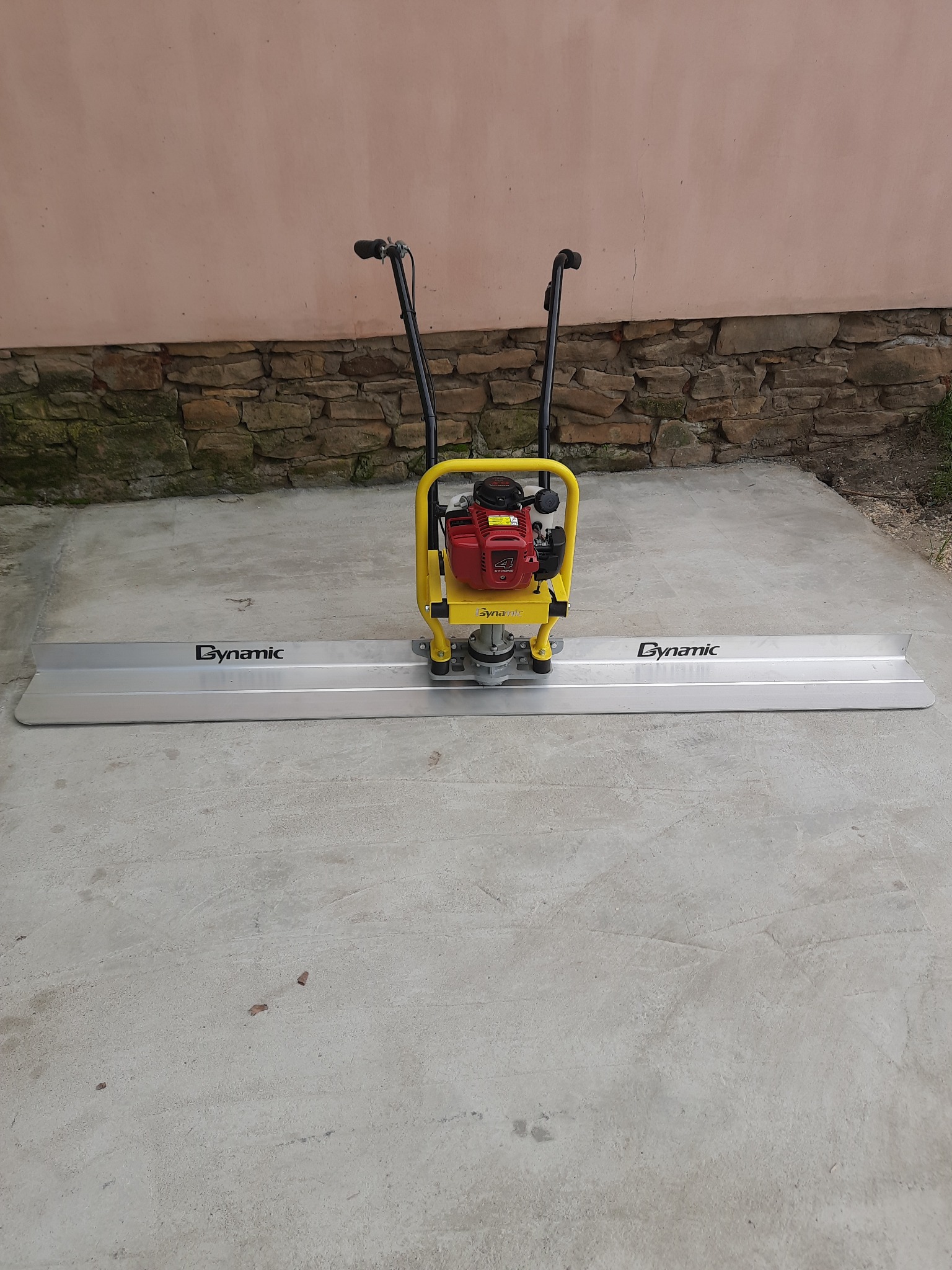 Listwa wibracyjna Dynamic do betonu z silnikiem spalinowym, żółto-czerwona, stojąca na szarym betonie, w tle mur z kamienia i różowa ściana.