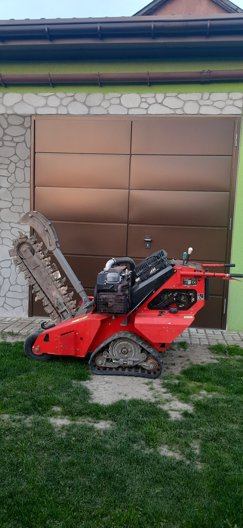 Czerwona glebogryzarka łańcuchowa na gąsienicach, zaparkowana na trawniku przed garażem z brązową bramą segmentową.