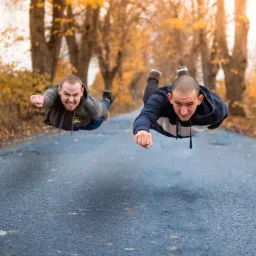Dwaj mężczyźni w bluzach z kapturem, udający lot superbohaterów na tle jesiennego krajobrazu z drzewami i opadłymi liśćmi na drodze asfaltowej.