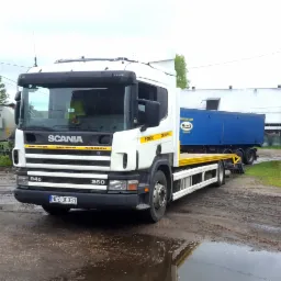 Biała laweta Scania 94D 260 na błotnistym podłożu, z niebieską naczepą w tle i żółtymi elementami platformy załadunkowej.