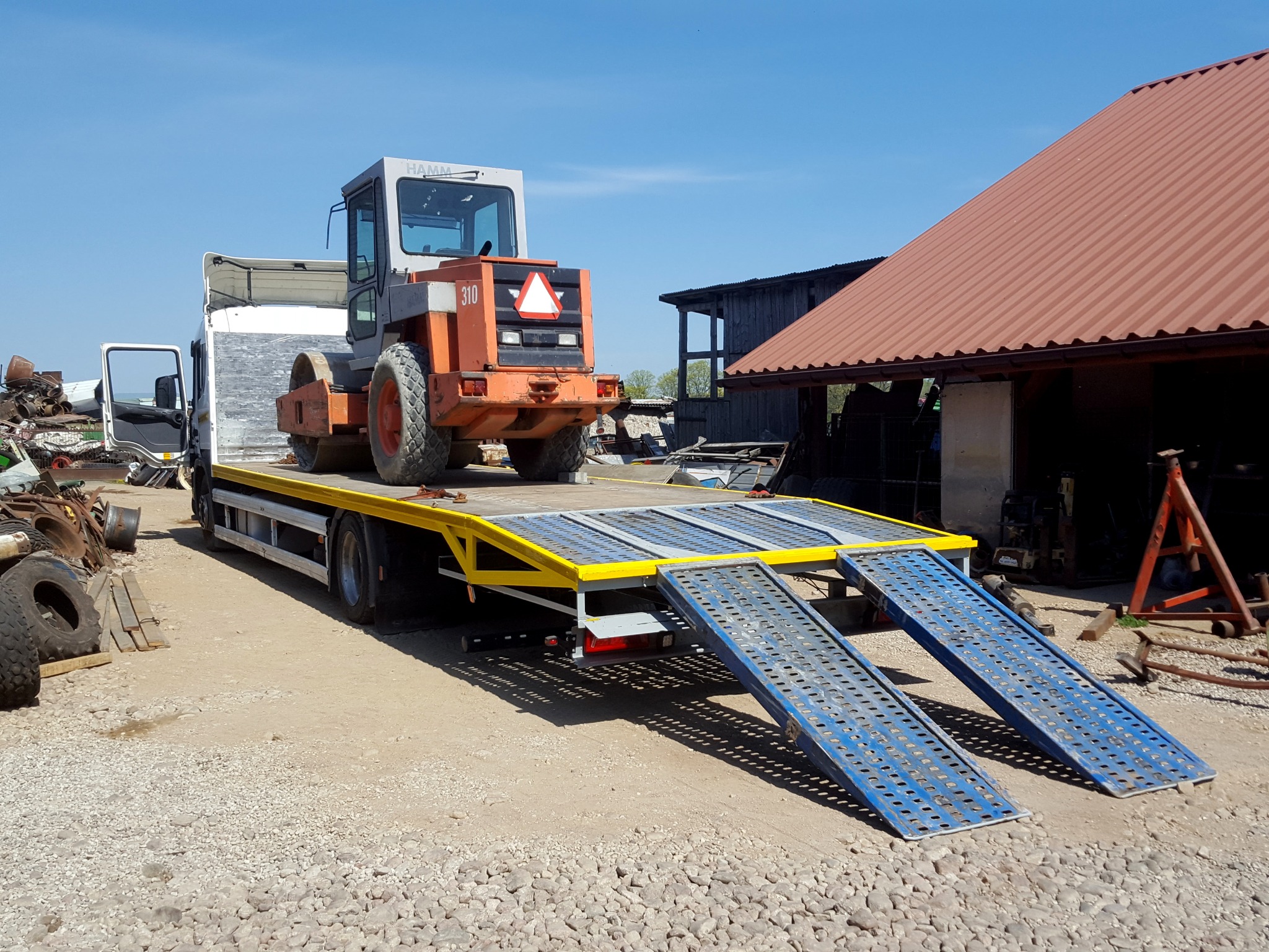 Laweta z Działdowa z pomarańczowym walcem drogowym marki Hamm na platformie, gotowa do transportu, z rampami opuszczonymi na żwirowe podłoże.