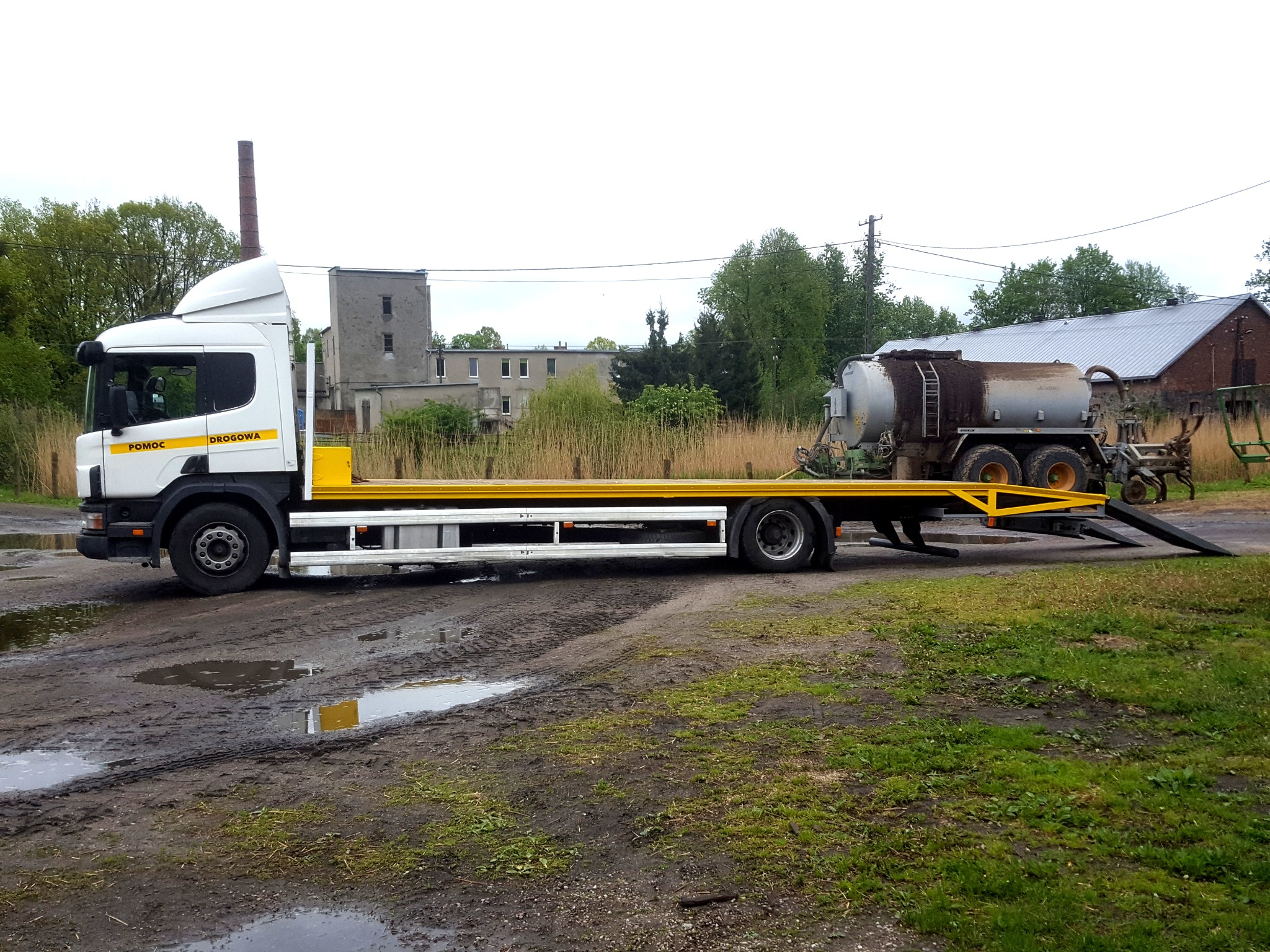 Biała ciężarówka z żółtymi elementami, oznaczona jako 'Pomoc Drogowa', przewozi brudną, srebrną cysternę rolniczą na platformie. W tle widać budynki i drzewa, a na pierwszym planie błotnisty teren...
