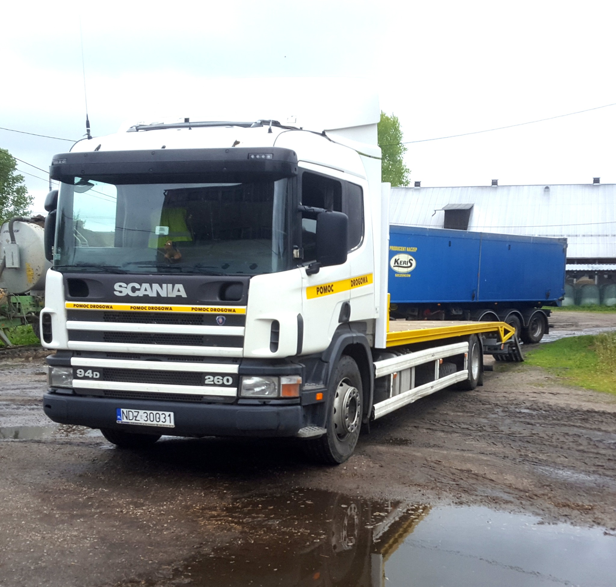 Biała laweta Scania 94D 260 na błotnistym podłożu, z niebieską naczepą w tle i żółtymi elementami platformy załadunkowej.