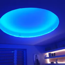 Okrągły podwieszany sufit z niebieskim oświetleniem LED, dodatkowo oświetlone półki w tle, widoczne rolety okienne.