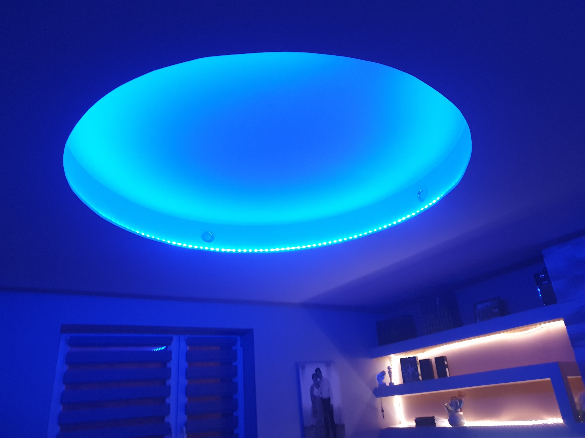 Okrągły podwieszany sufit z niebieskim oświetleniem LED, dodatkowo oświetlone półki w tle, widoczne rolety okienne.