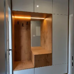 Biało-drewniana szafa wnękowa z oświetleniem LED i wieszakami na ubrania w przedpokoju, widok z perspektywy korytarza.