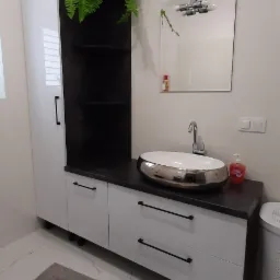 Nowoczesna łazienka z białą szafką pod umywalkę z czarnymi uchwytami, umywalką nablatową z chromowanymi brzegami, lustrem z oświetleniem i zieloną paprotką na szafce.