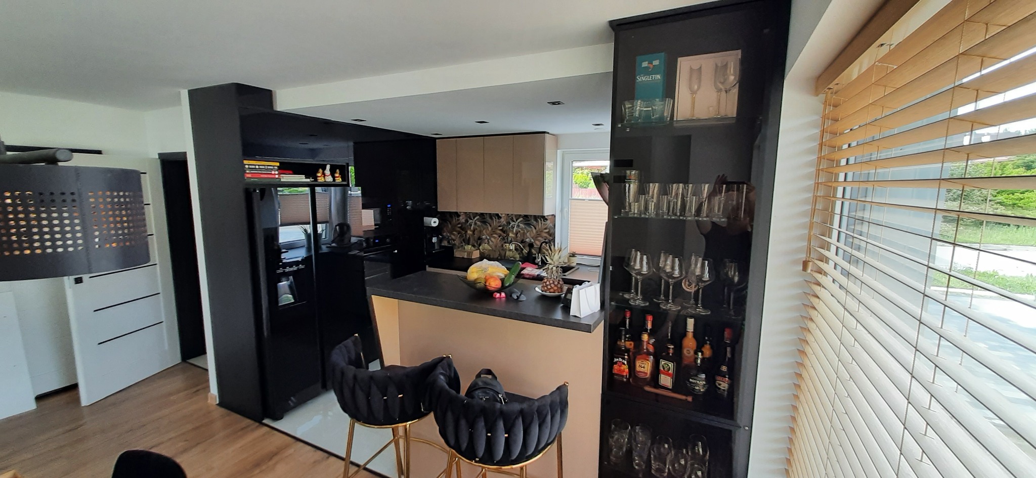Nowoczesna kuchnia z barkiem, czarnymi krzesłami barowymi na złotych nogach, przeszkloną szafką z alkoholem i owocami na blacie.