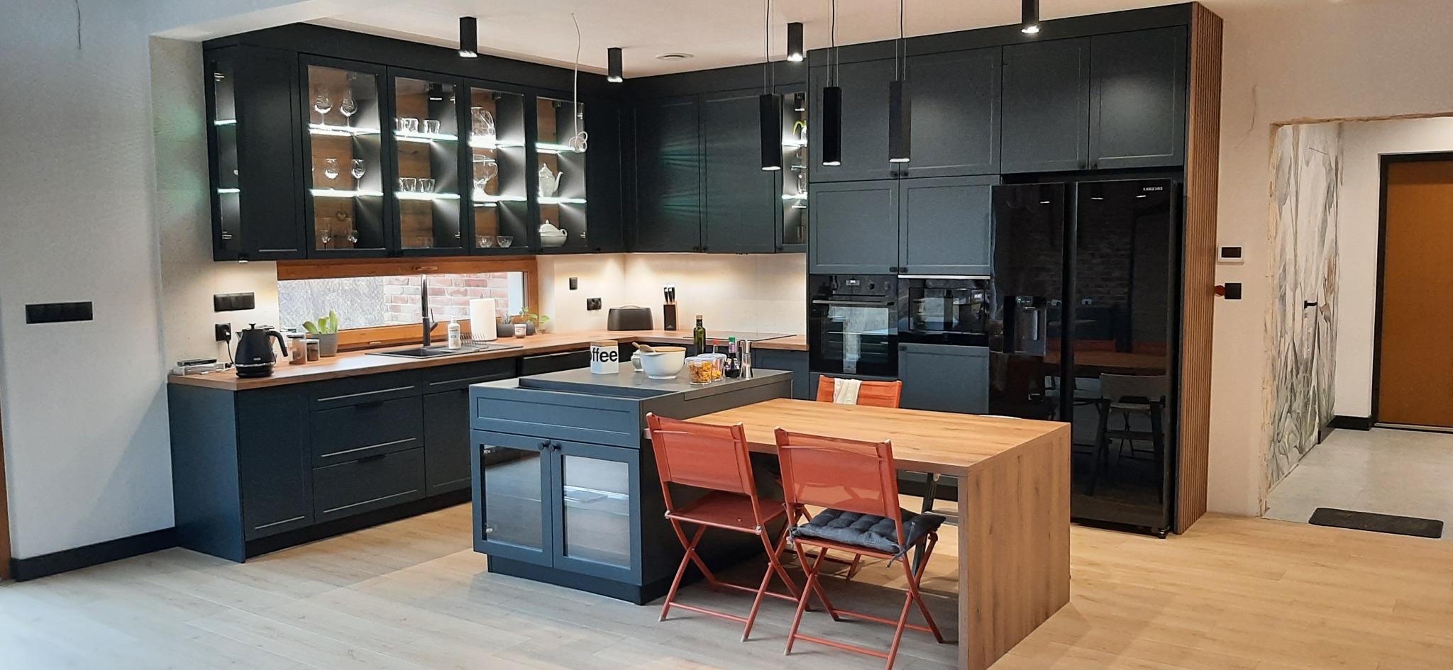 Nowoczesna kuchnia w odcieniach grafitu z drewnianymi blatami i wyspą, oświetlona wiszącymi lampami, z przeszklonymi szafkami górnymi podświetlonymi LED, z widocznym stołem i krzesłami.