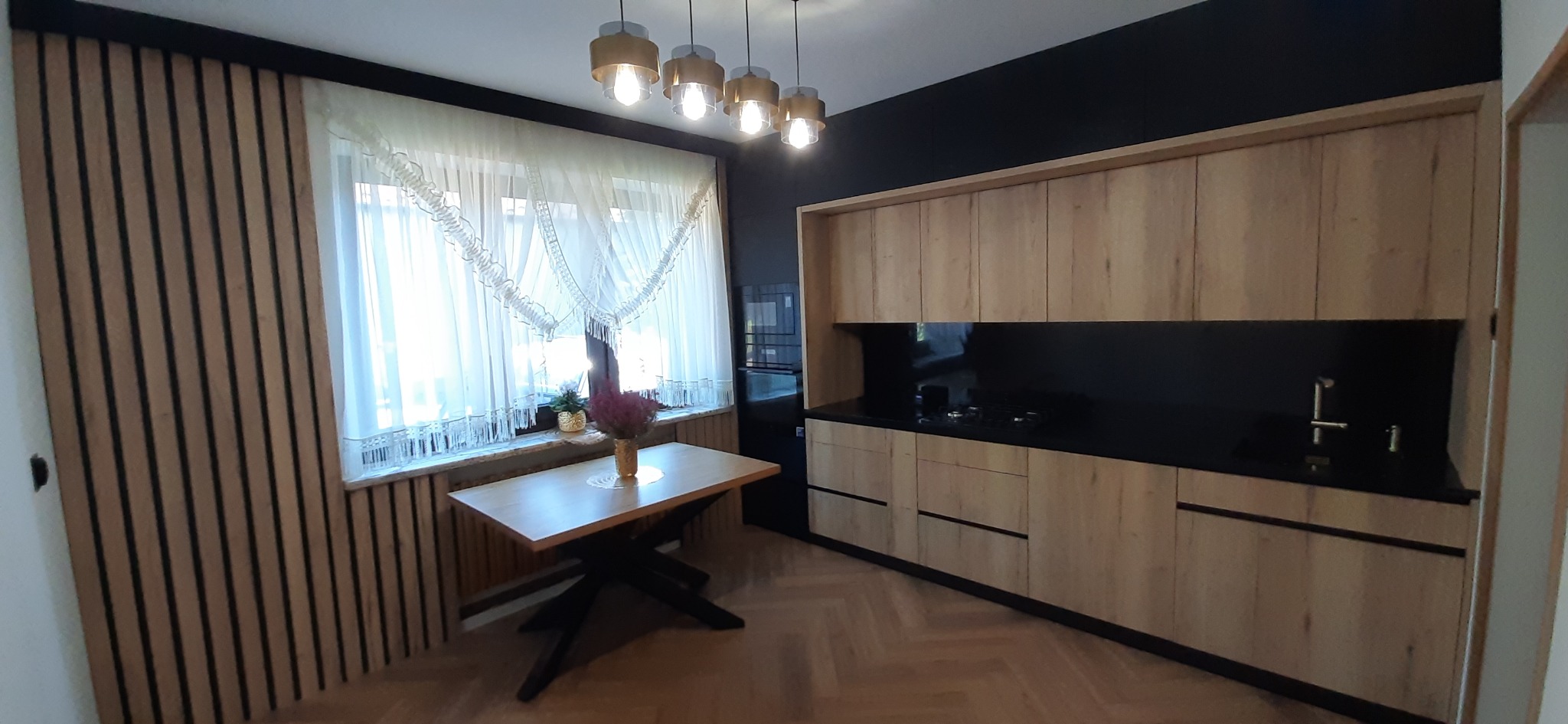 Jasna kuchnia z drewnianymi frontami i czarnym blatem, uzupełniona o stół z kwiatami i dekoracyjne oświetlenie.