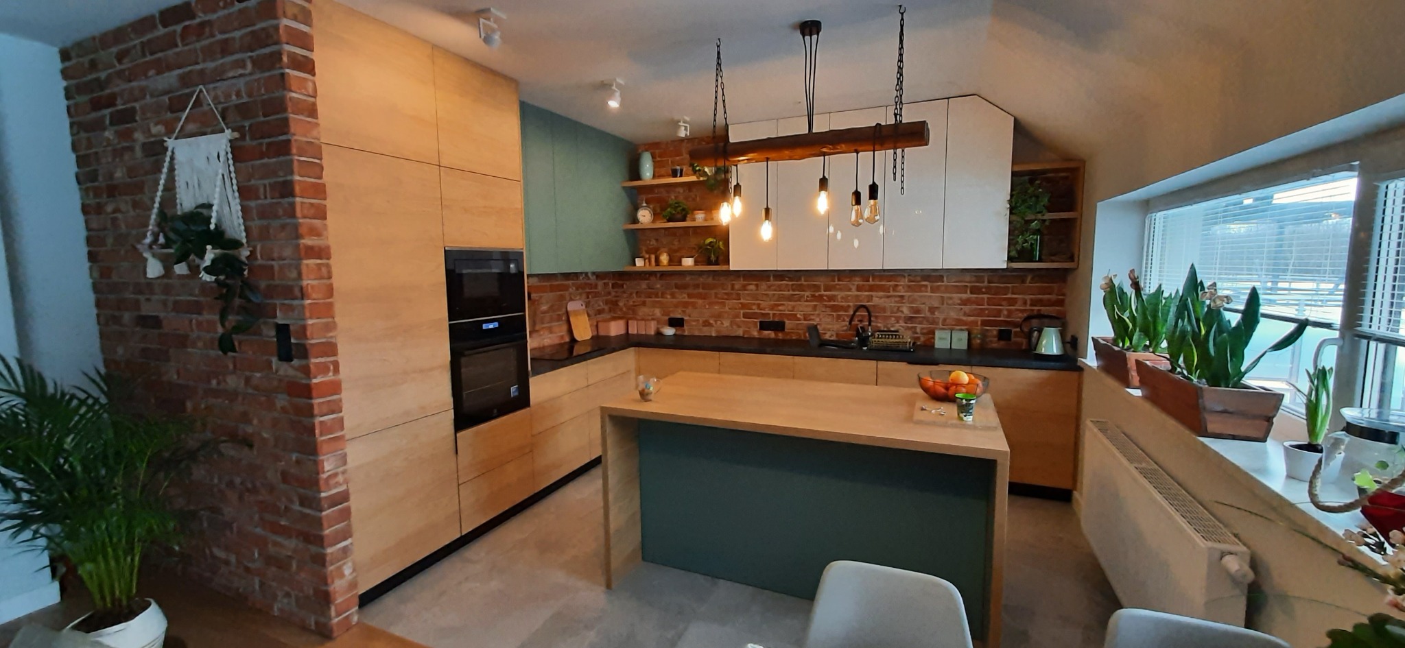 Nowoczesna kuchnia z wyspą, drewnianymi frontami, ceglaną ścianą i wiszącymi lampami z żarówkami Edisona.