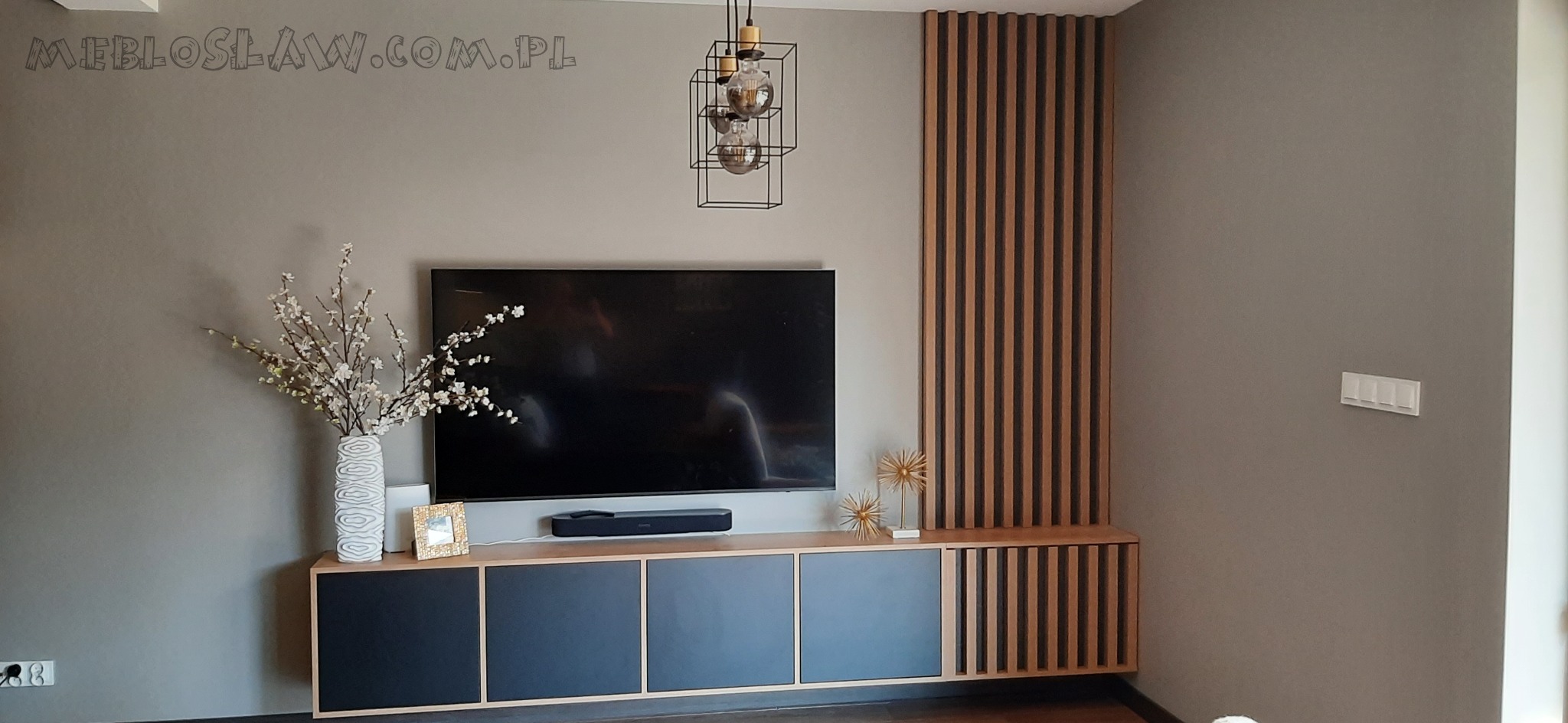 Nowoczesna szafka RTV z czarnymi frontami, drewnianym blatem i dekoracyjną ścianką z drewnianych listewek, uzupełniona o wazon z kwiatami i minimalistyczną lampę wiszącą.