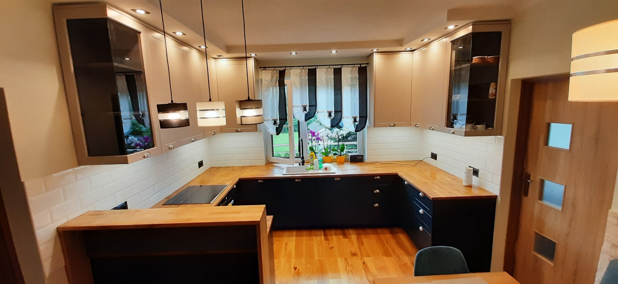 U-kształtna kuchnia z drewnianym blatem, czarnymi dolnymi szafkami i beżowymi górnymi szafkami z przeszklonymi witrynami, oświetlona trzema wiszącymi lampami i punktowymi światłami, z widokiem...