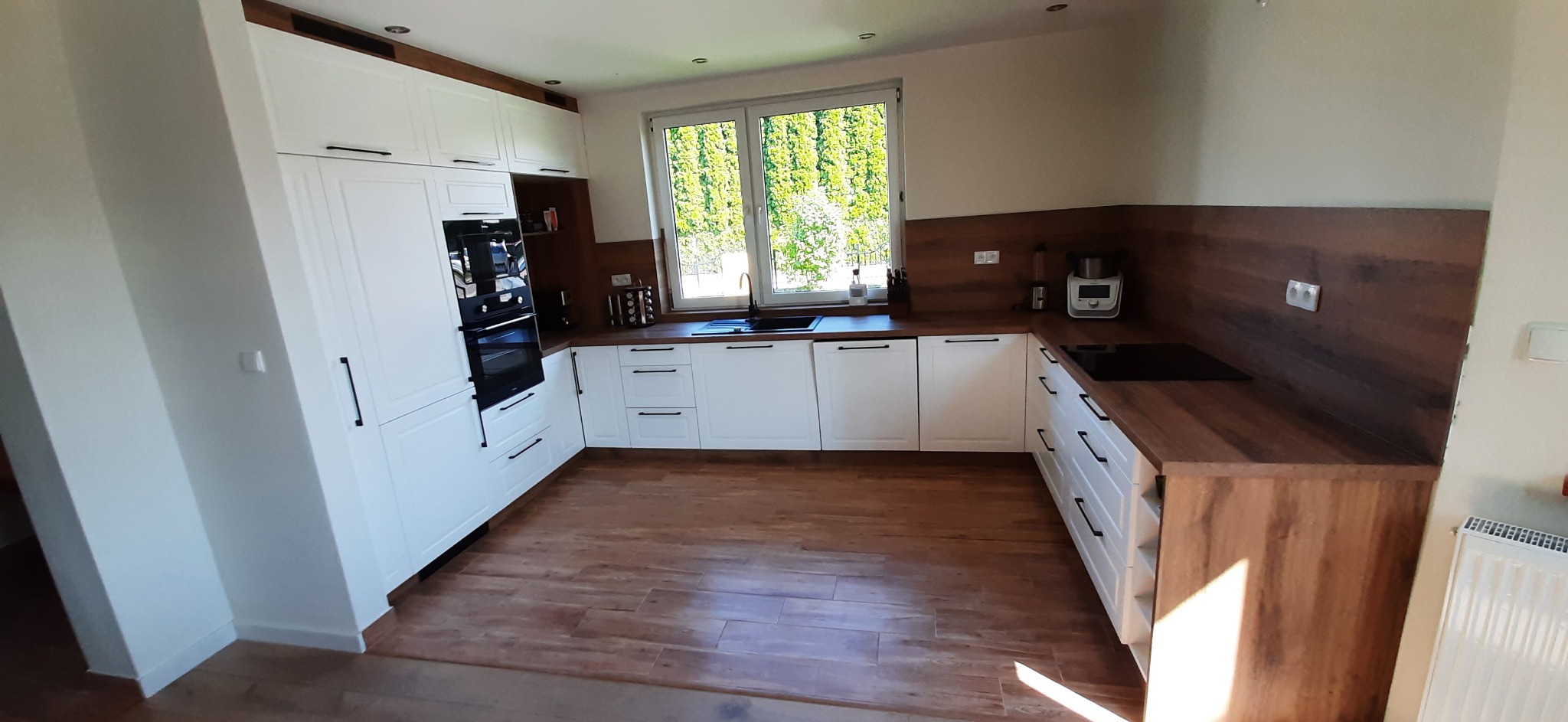 Jasna kuchnia z białymi szafkami i drewnianym blatem, czarne uchwyty, okno z widokiem na zieleń, drewniana podłoga z płytek.