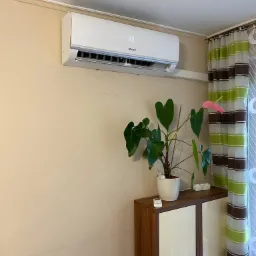 Biały klimatyzator Mistral z wyświetlaczem temperatury 23 stopnie Celsjusza, zamontowany na ścianie w pomieszczeniu w Krakowie, obok szafki z kwiatem anturium i zasłonami w paski.