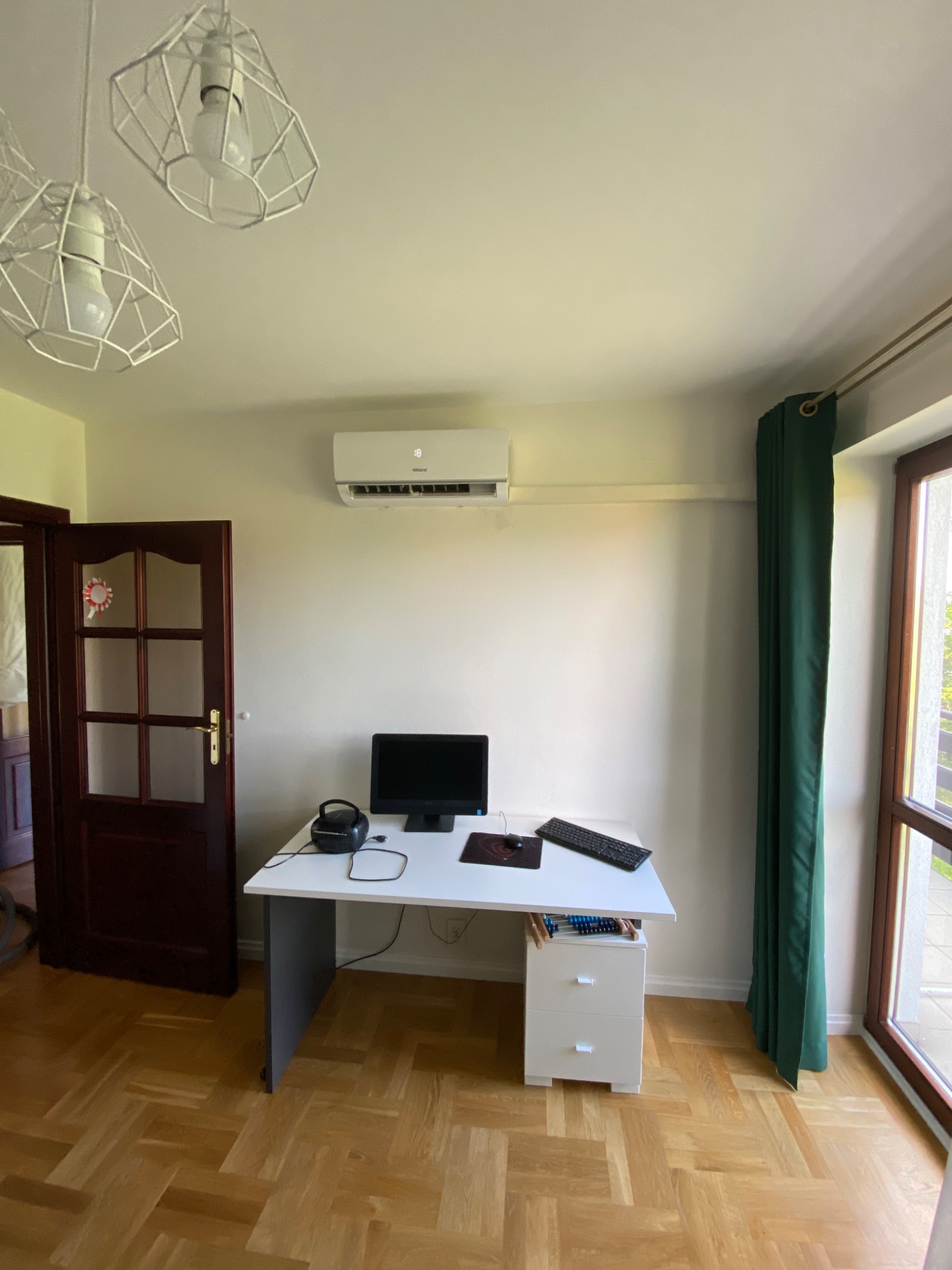 Biały klimatyzator ścienny zamontowany w pokoju nad biurkiem z komputerem, oświetlonym przez designerskie lampy wiszące z metalowymi kloszami.