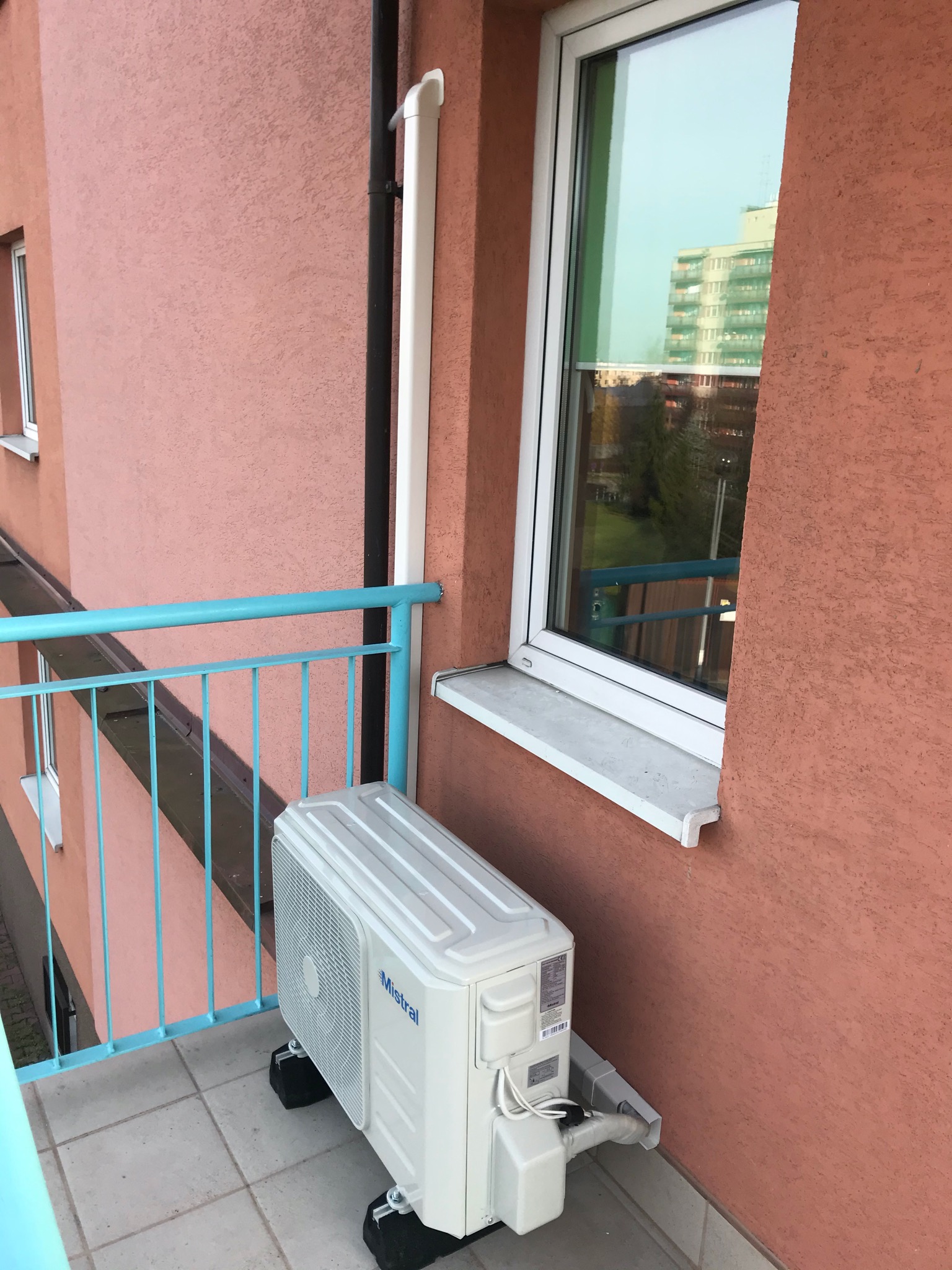 Zewnętrzna jednostka klimatyzatora Mistral 3,5kW zamontowana na balkonie bloku mieszkalnego, widoczna turkusowa balustrada balkonu i odbicie budynku w oknie.