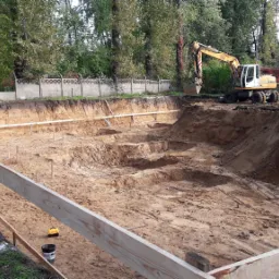 Żółty traktor kopie dół w ziemi, widoczne drewniane wzmocnienia i ogrodzenie, w tle drzewa i budynki.