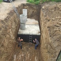 Trans-Kop - Głęboki wykop z betonową konstrukcją i dwoma pracownikami w środku, widoczne betonowe kręgi, drabiny, brązowy samochód osobowy i ziemia.