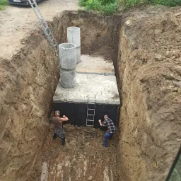 Głęboki wykop z betonową konstrukcją i dwoma pracownikami w środku, widoczne betonowe kręgi, drabiny, brązowy samochód osobowy i ziemia.