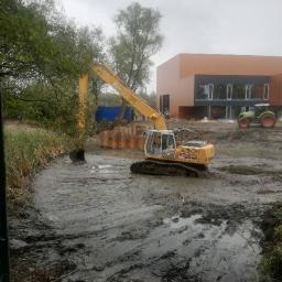 Trans-Kop - Żółta koparka Liebherr 904C pracuje w błotnistym wykopie, w tle budynek z brązową elewacją i zielony traktor, pochmurne niebo.