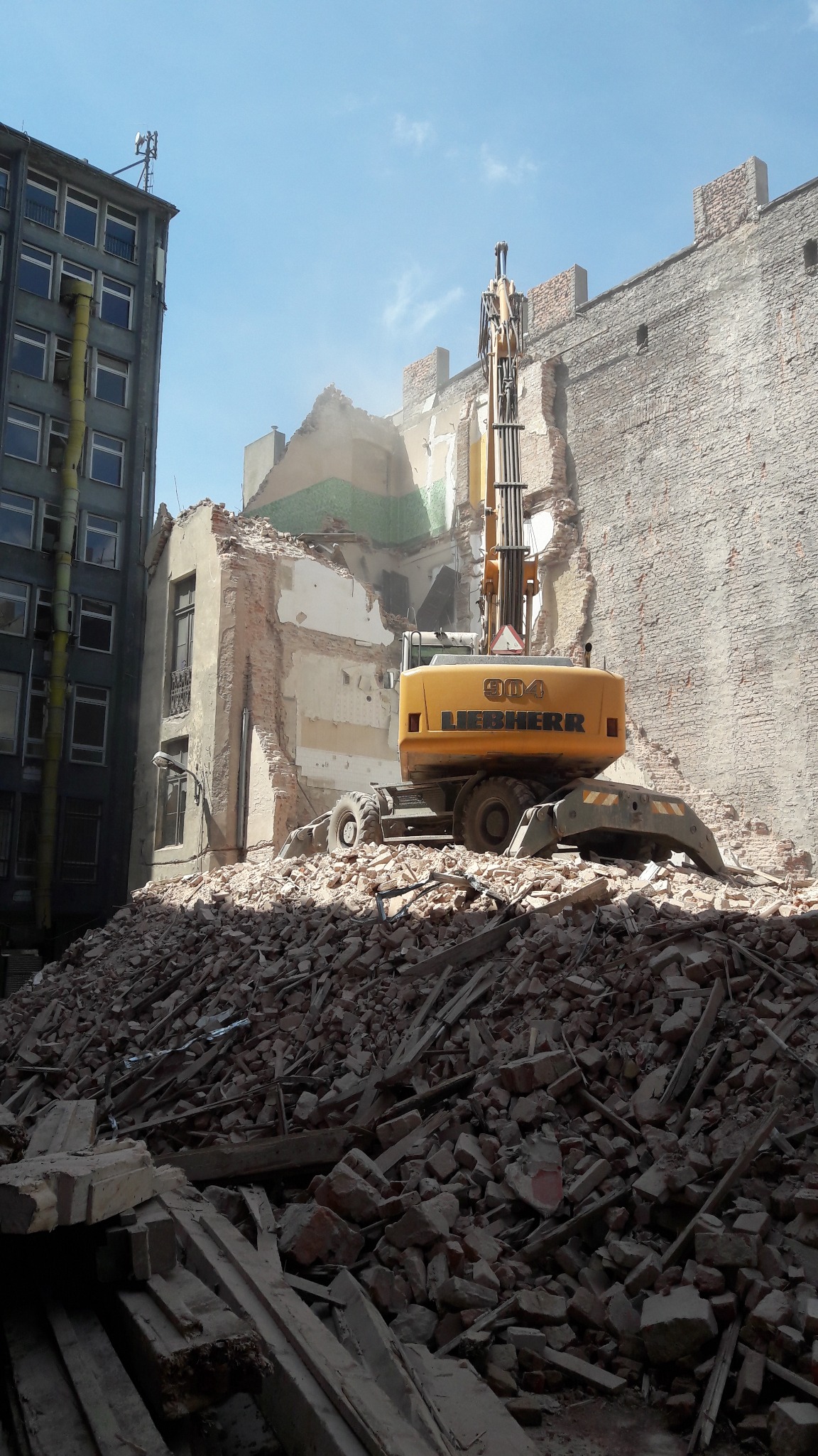 Żółta maszyna Liebherr 904 na stercie gruzu podczas rozbiórki budynku w mieście, z widocznym fragmentem ściany sąsiedniego budynku.