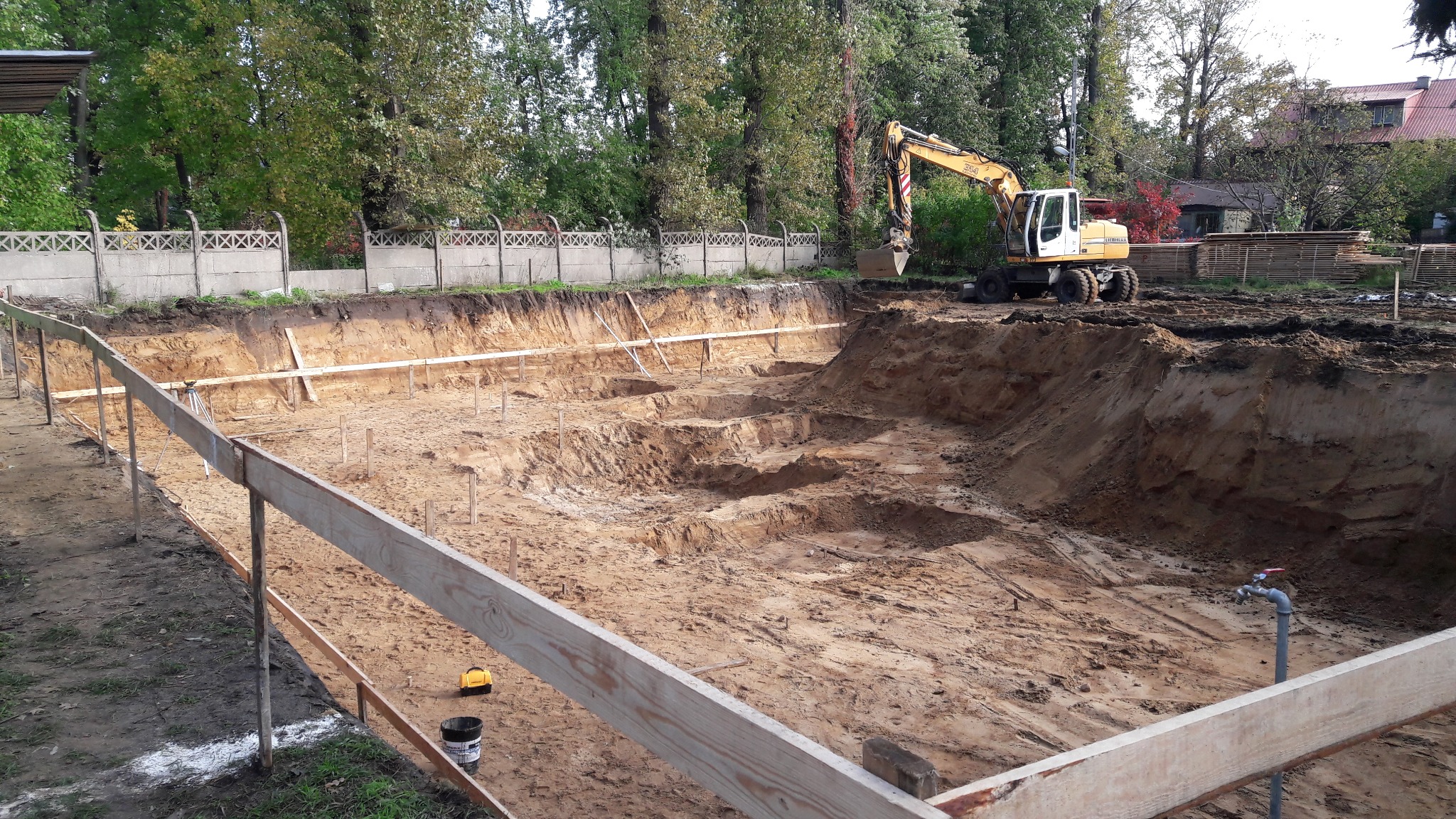 Żółty traktor kopie dół w ziemi, widoczne drewniane wzmocnienia i ogrodzenie, w tle drzewa i budynki.