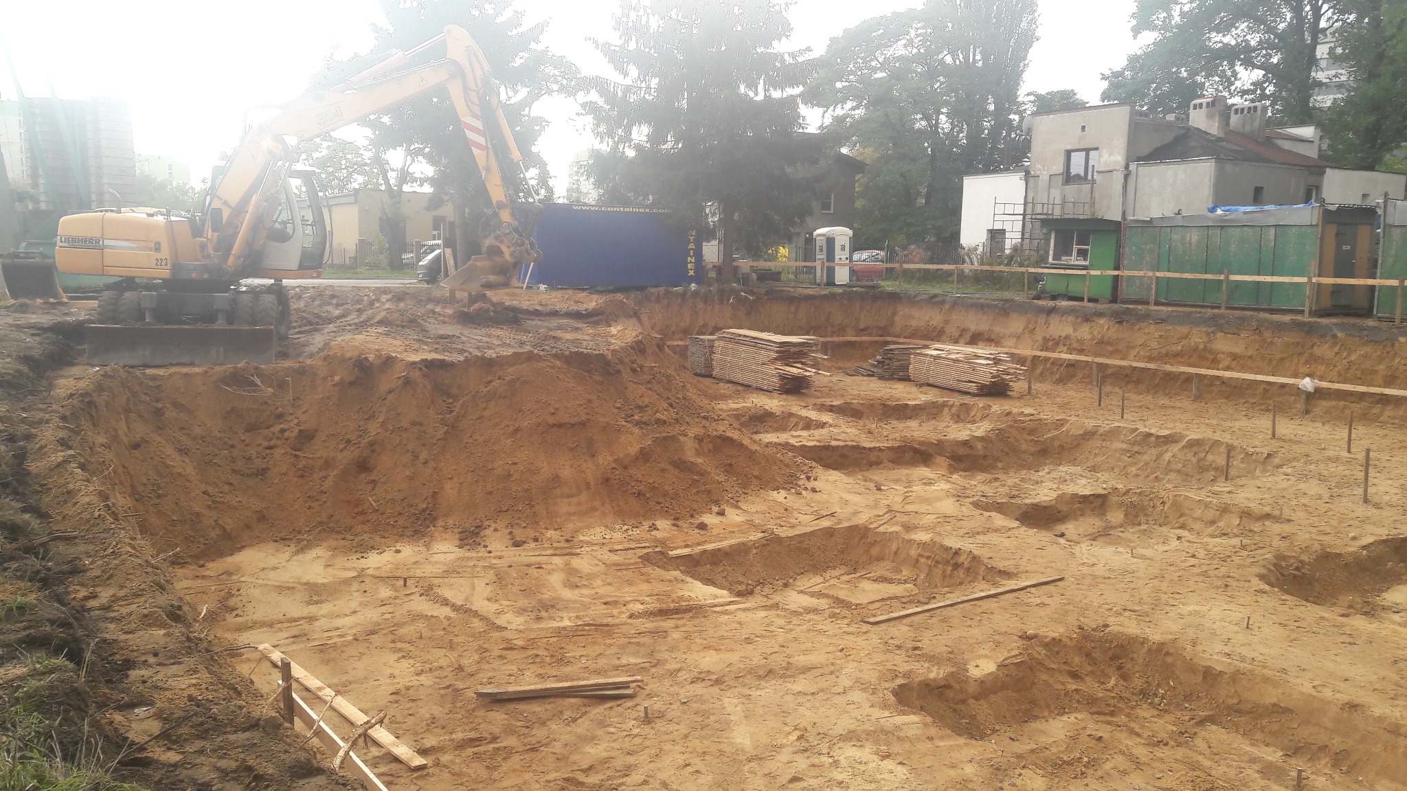 Żółty koparka Liebherr 223 na skraju wykopu pod budynek z widocznymi stertami drewna i zabezpieczeniami brzegów.