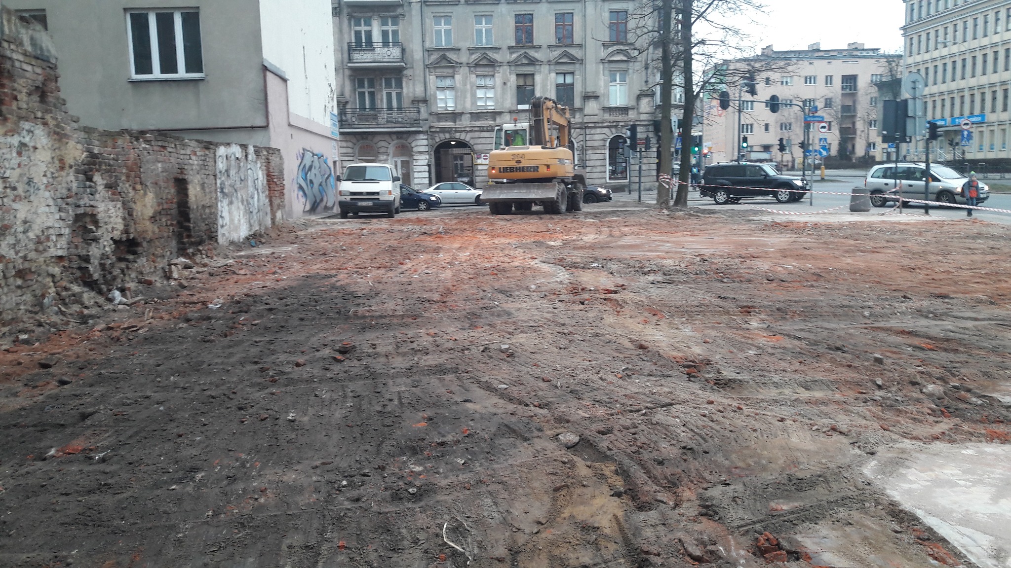 Żółta koparka Liebherr na placu budowy z częściowo rozebranym murem z cegły w tle, widoczne ślady opon na ziemi, miejski krajobraz z budynkami i samochodami.