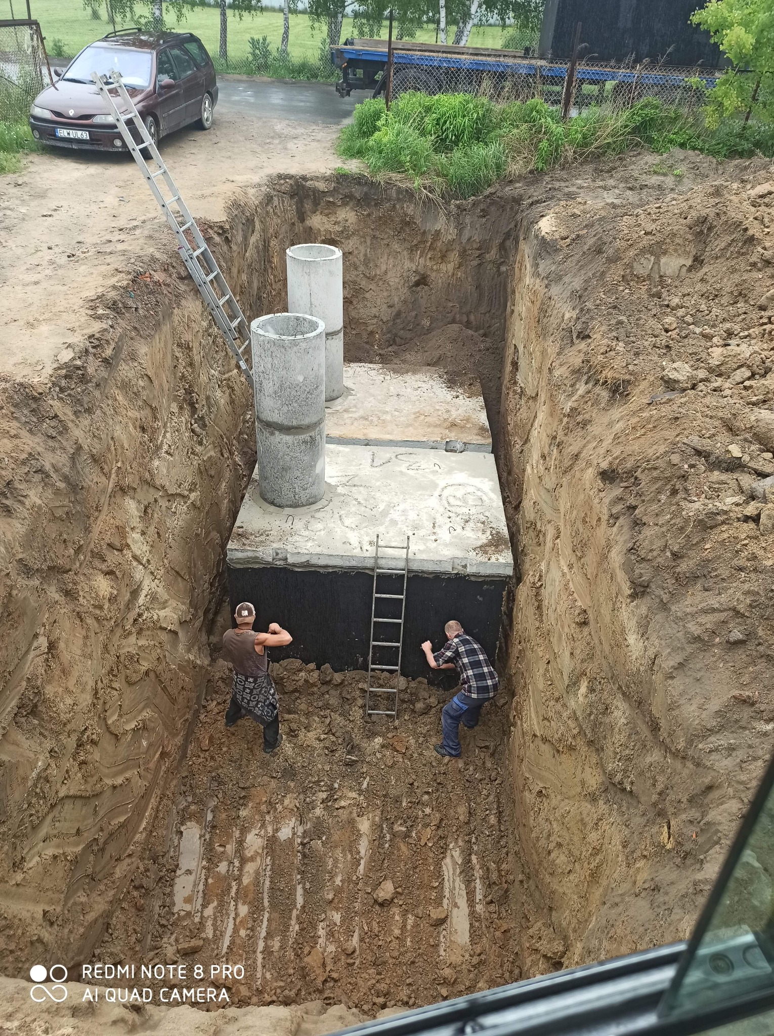 Głęboki wykop z betonową konstrukcją i dwoma pracownikami w środku, widoczne betonowe kręgi, drabiny, brązowy samochód osobowy i ziemia.