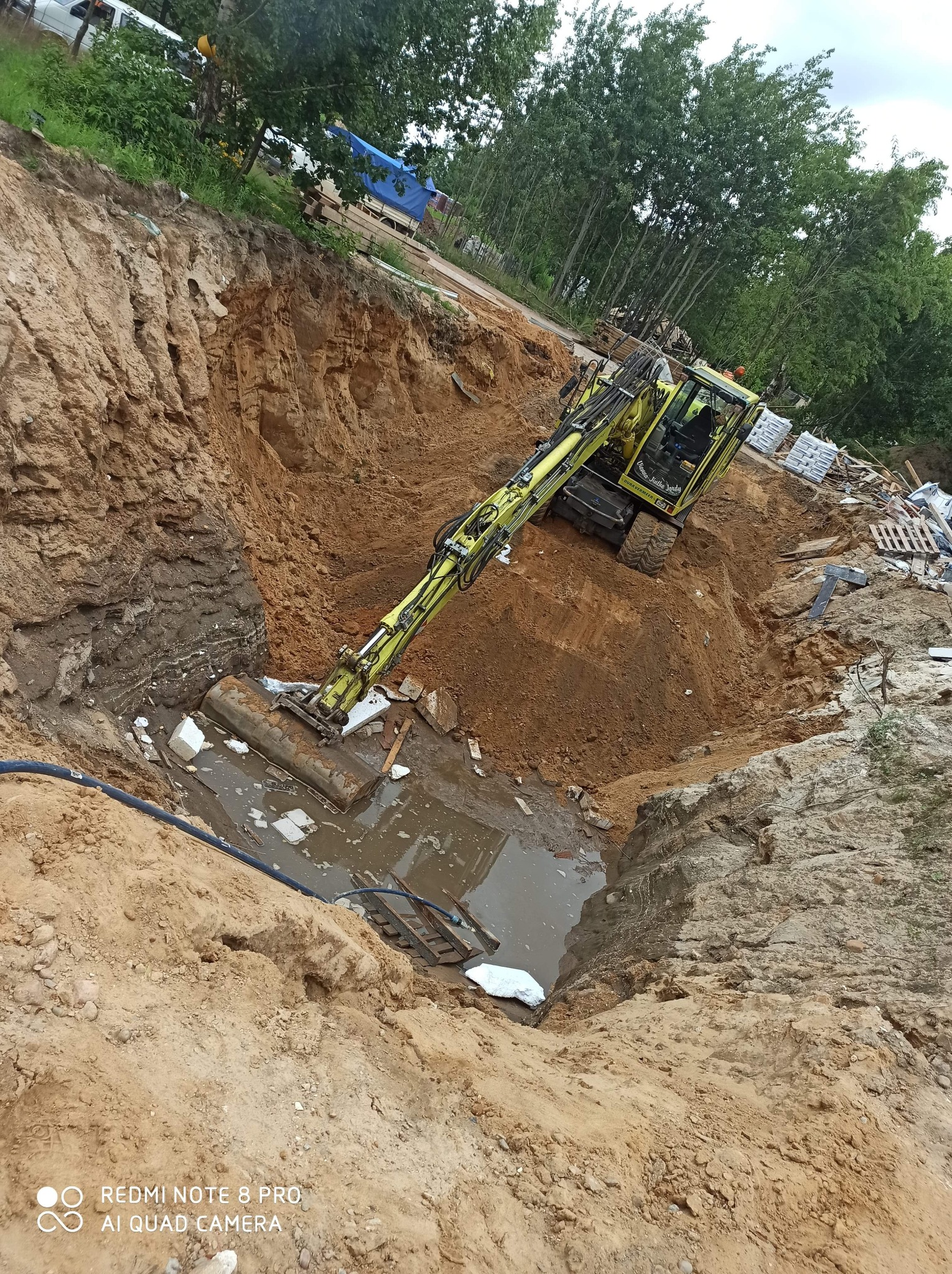 Żółta koparka w głębokim wykopie z wodą i elementami konstrukcyjnymi, widok z góry.