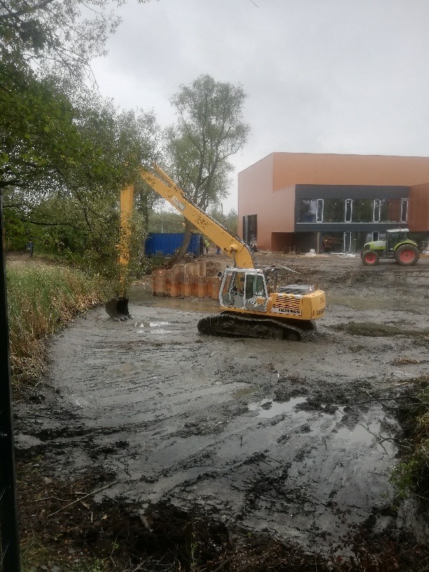 Żółta koparka Liebherr 904C pracuje w błotnistym wykopie, w tle budynek z brązową elewacją i zielony traktor, pochmurne niebo.