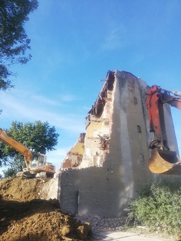 Dwa pomarańczowe i żółte koparki pracują przy częściowo zburzonym, ceglanym budynku pod błękitnym niebem. Widoczne odsłonięte fragmenty ścian i gruz.