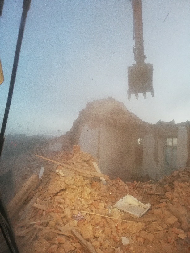 Demolka budynku mieszkalnego z użyciem koparki, widok z kabiny operatora, sterta gruzu ceglanego i drewnianych elementów konstrukcyjnych