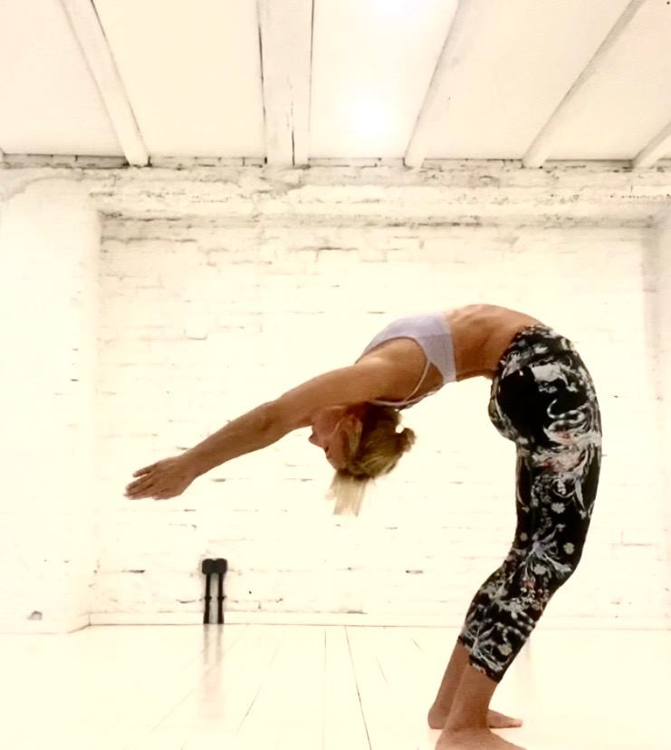 Kobieta w sportowym stroju wykonuje mostek w jasnym pomieszczeniu z ceglaną ścianą i drewnianym sufitem, widoczne czarne wałki do masażu.