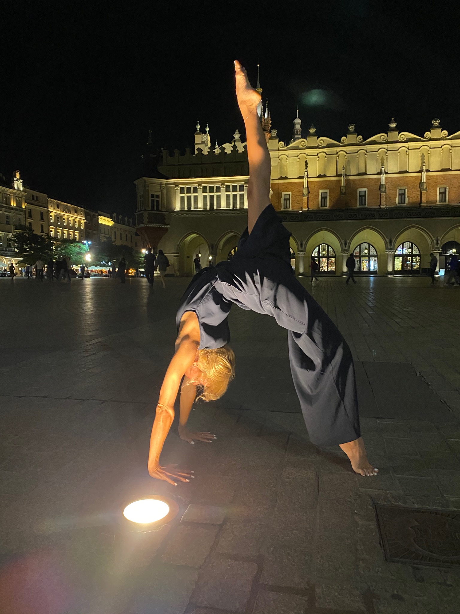 Kobieta w kombinezonie wykonuje pozycję mostka z uniesioną nogą na tle Sukiennic w Krakowie, oświetlona lampą podłogową.
