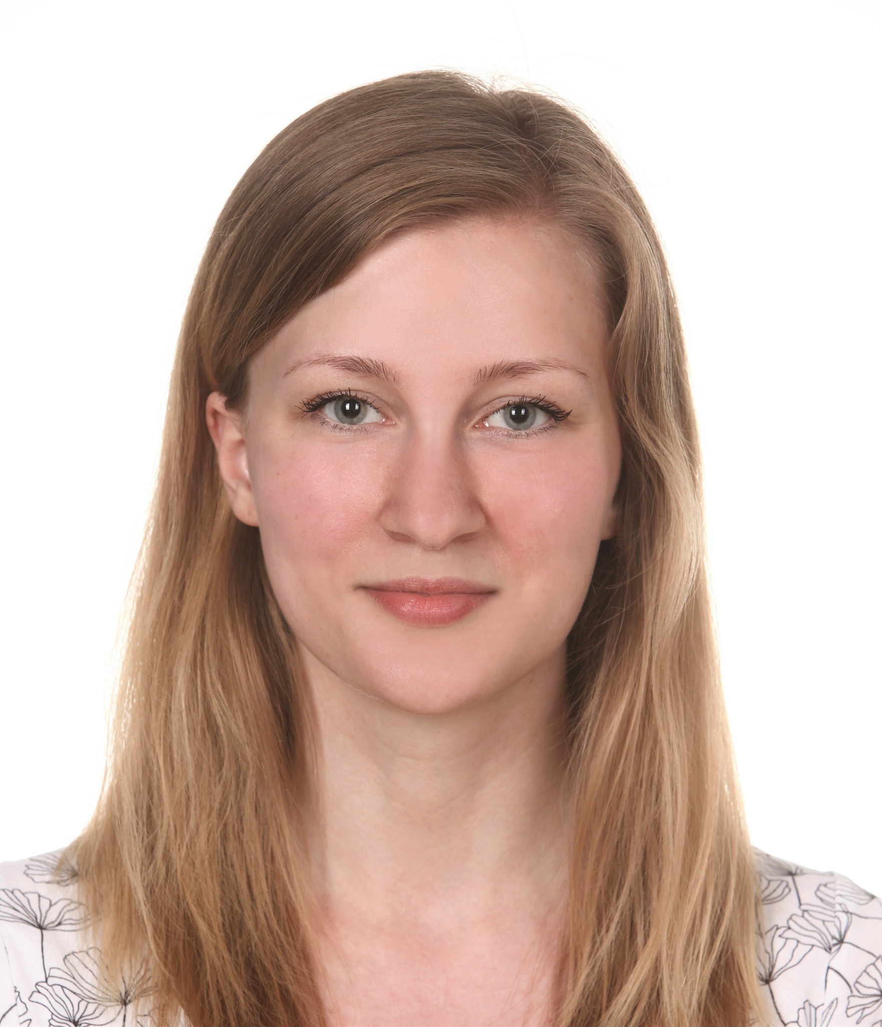 Portret młodej kobiety z blond włosami i jasną cerą, uśmiechająca się delikatnie, ubrana w białą bluzkę z czarnymi kwiatami na tle białego studia.