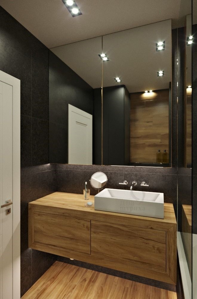 Nowoczesna łazienka z drewnianą szafką pod umywalką, prostokątną białą umywalką nablatową, dużym lustrem i czarnymi płytkami na ścianie. Widoczne oświetlenie sufitowe i podświetlenie lustra.